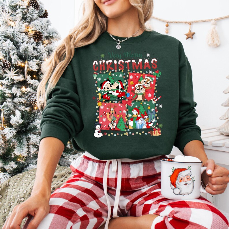 May include: A dark green sweatshirt with a festive Christmas design featuring cartoon characters. The words "Very Merry Christmas" are displayed above the graphic. The person is wearing red and white plaid pajama pants and holding a mug with a Santa Claus design.