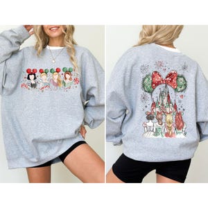 May include: A light gray sweatshirt featuring a festive design. The front displays four cartoon characters with holiday-themed mouse ears. The back showcases a castle scene with a large red bow and mouse ears. The sweatshirt is paired with black shorts.