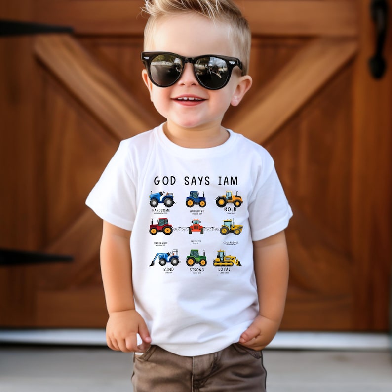 May include: White t-shirt with the text "God Says I Am" and a graphic of tractors with words like "handsome", "accepted", "bold", "blessed", "kind", "strong", and "loyal" underneath each tractor.