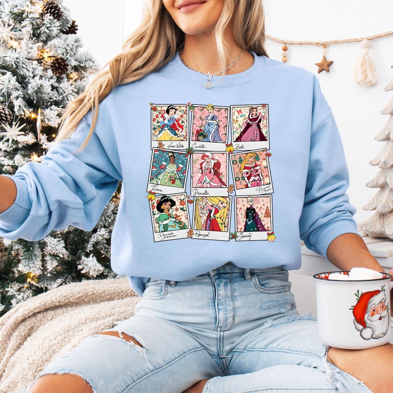 Puede incluir: Sudadera azul claro con nueve retratos de princesas en marcos estilo polaroid. Cada marco tiene el nombre de una princesa. La sudadera es de cuello redondo. El tema general son las princesas de Disney.