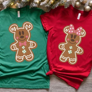 May include: Two green and red t-shirts featuring a gingerbread Mickey Mouse and Minnie Mouse design. The Mickey Mouse shirt is green and the Minnie Mouse shirt is red.