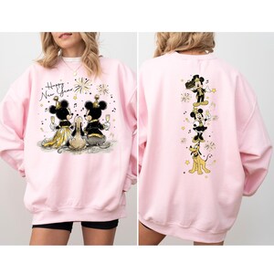 Puede incluir: Sudadera rosa claro con un dise&ntilde;o festivo de Mickey y Minnie Mouse celebrando el A&ntilde;o Nuevo. La parte delantera muestra a la pareja con copas de champ&aacute;n, mientras que la parte trasera los muestra con Pluto. Tambi&eacute;n se ve el texto "Happy New Year".
