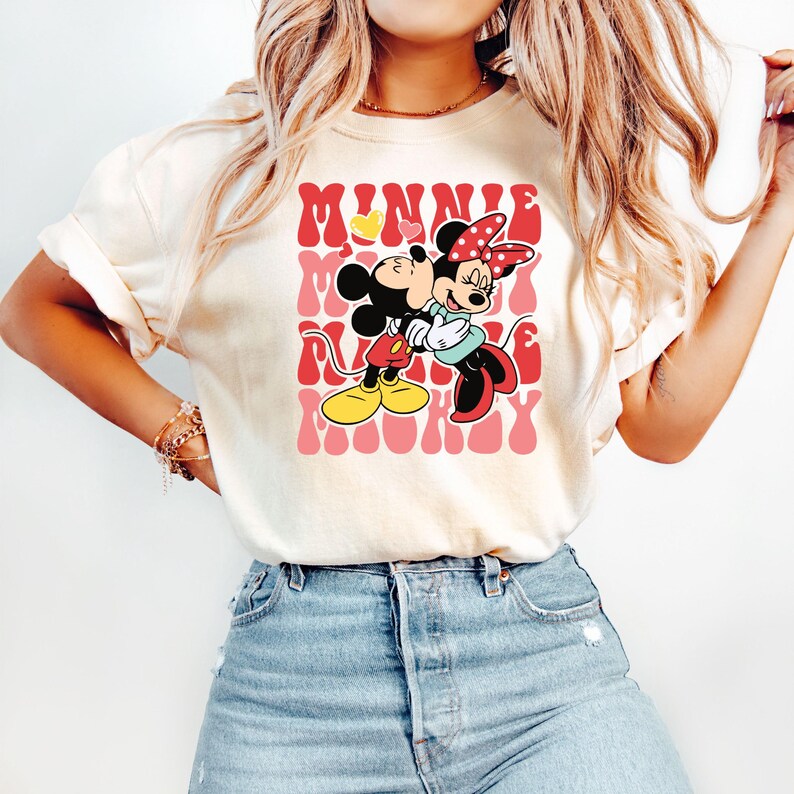 K&ouml;nnte beinhalten: Cremefarbenes T-Shirt mit einer Grafik von Mickey und Minnie Mouse, die sich umarmen, mit dem Wort "MINNIE" in roter Retro-Schrift. Das Shirt hat eine lockere Passform und kurze &Auml;rmel. Das Design enth&auml;lt gelbe Herzen.