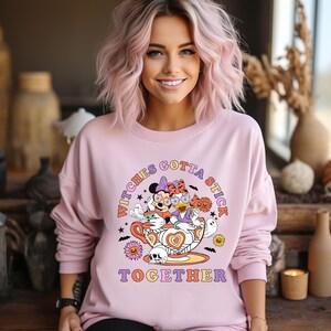 K&ouml;nnte beinhalten: Ein rosa Sweatshirt mit einem Aufdruck von Mickey Mouse und Minnie Mouse, die als Hexen in einer Teetasse verkleidet sind. Der Text "Witches Gotta Stick Together" ist auf dem Sweatshirt aufgedruckt.