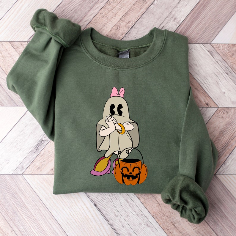Puede incluir: Una sudadera verde con un fantasma blanco que lleva un lazo rosa y sostiene un cubo de calabaza. El fantasma tiene una expresi&oacute;n de sorpresa con los ojos muy abiertos.