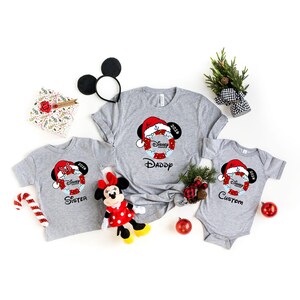 K&ouml;nnte beinhalten: Graue Familien-Weihnachtsshirts mit einem Disney Mickey Mouse-Design mit Weihnachtsm&uuml;tze. Die Shirts sind mit "Daddy", "Sister" und "Custom" beschriftet. Ein Minnie Mouse-Pl&uuml;schtier, eine Zuckerstange und Weihnachtsdekorationen sind ebenfalls vorhanden.