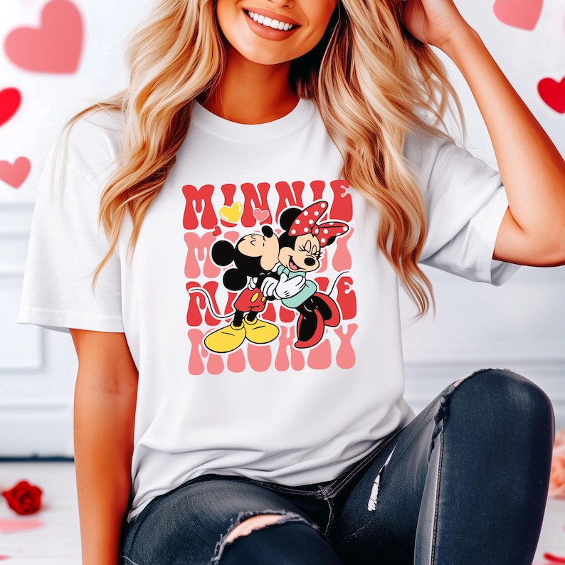 K&ouml;nnte beinhalten: Wei&szlig;es T-Shirt mit einer Cartoon-Illustration von Mickey und Minnie Mouse, die sich umarmen. Das Design enth&auml;lt die Worte "Minnie Mickey" in einem sich wiederholenden Muster mit roten und rosa Farbt&ouml;nen. Die Charaktere tragen ihre klassischen Outfits.