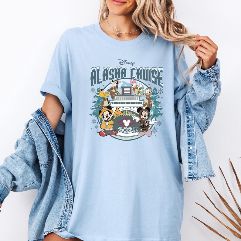 Pu&ograve; includere: T-shirt azzurra con un design Disney Alaska Cruise. La grafica include Topolino e Minnie, Pippo, Paperino e Paperina, una nave da crociera e l'anno 2025. Le maniche sono arrotolate.