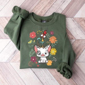 Puede incluir: Una sudadera verde con una imagen de dibujos animados de un cerdo y un gallo con detalles florales. El cerdo es blanco con manchas negras y el gallo es rojo, amarillo y verde.