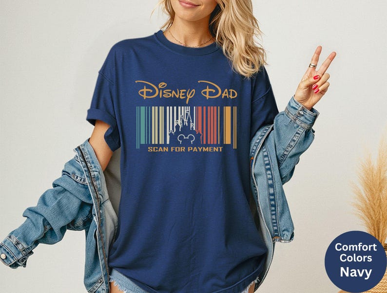 May include: Navy blue t-shirt featuring the text "Disney Dad" in gold script above a barcode design. The barcode includes a castle silhouette and Mickey Mouse ears. The text "SCAN FOR PAYMENT" is below.