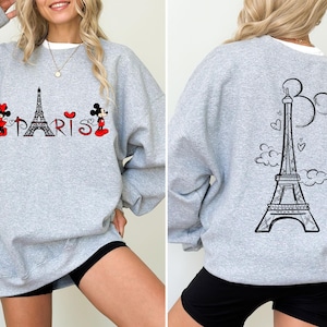 K&ouml;nnte beinhalten: Ein graues Sweatshirt mit einem Design mit dem Eiffelturm und dem Wort "Paris". Die Vorderseite des Sweatshirts zeigt Minnie- und Mickey Mouse-Figuren. Die R&uuml;ckseite des Sweatshirts hat eine schwarze Umrisszeichnung des Eiffelturms.