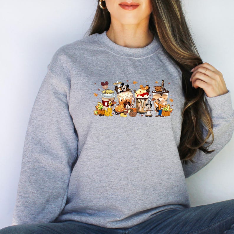 May include: A gray sweatshirt featuring a colorful graphic of cartoon characters in coffee cups with fall-themed decorations. The design includes pumpkins, leaves, and various characters, creating a festive autumn theme.
