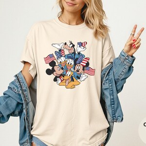 May include: Ivory-colored t-shirt featuring a graphic of Mickey Mouse, Minnie Mouse, Donald Duck, Daisy Duck, Goofy, and Pluto, each holding or wearing American flags. The shirt is a comfort color and is ivory in color.