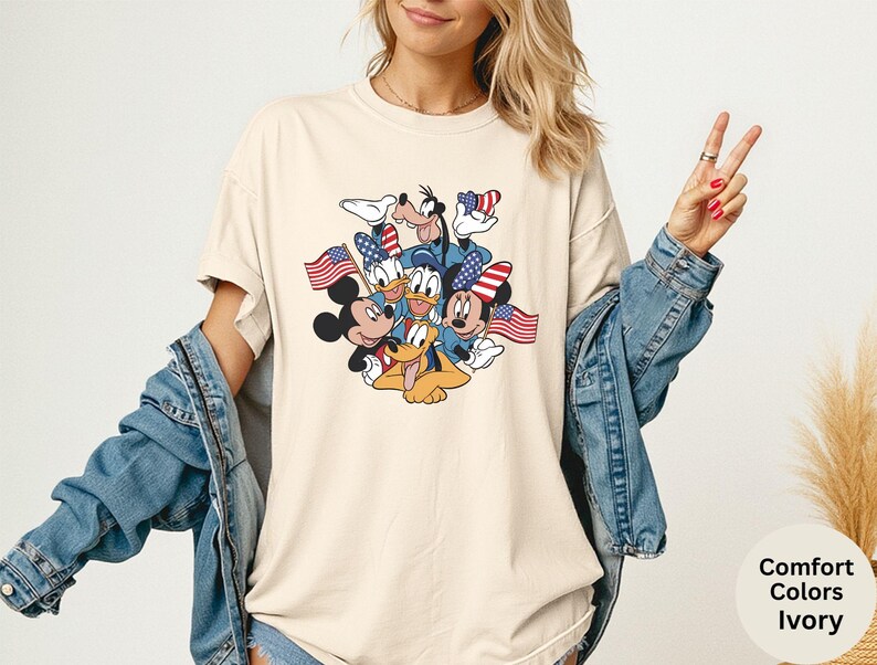 May include: Ivory-colored t-shirt featuring a graphic of Mickey Mouse, Minnie Mouse, Donald Duck, Daisy Duck, Goofy, and Pluto, each holding or wearing American flags. The shirt is a comfort color and is ivory in color.