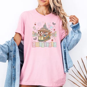 May include: Pink t-shirt featuring a Disneyland design. The graphic includes a trolley with Mickey Mouse, Donald Duck, and Goofy, with the word "Disneyland" below. The design is in pastel colors with fireworks and balloons.