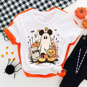 May include: White t-shirt with a vintage-style illustration of Mickey Mouse dressed as a ghost holding a jack-o-lantern. The illustration includes a tombstone that says "RIP" and other Halloween imagery.