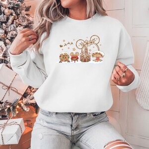 May include: White sweatshirt featuring a festive gingerbread design. The design includes gingerbread houses, a mouse-shaped head, and a reindeer-pulled sleigh. The sweatshirt is a comfortable, casual style, perfect for the holiday season.