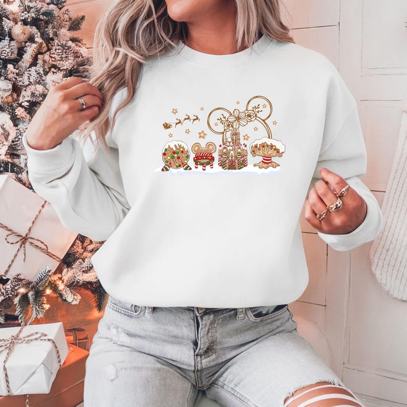 May include: White sweatshirt featuring a festive gingerbread design. The design includes gingerbread houses, a mouse-shaped head, and a reindeer-pulled sleigh. The sweatshirt is a comfortable, casual style, perfect for the holiday season.