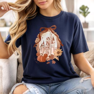 May include: Navy blue t-shirt with a castle illustration inside a brown frame, decorated with a bow, pumpkin, and autumn leaves. The design includes a sunflower and leaves, creating a seasonal theme. The shirt is a comfortable, casual wear.