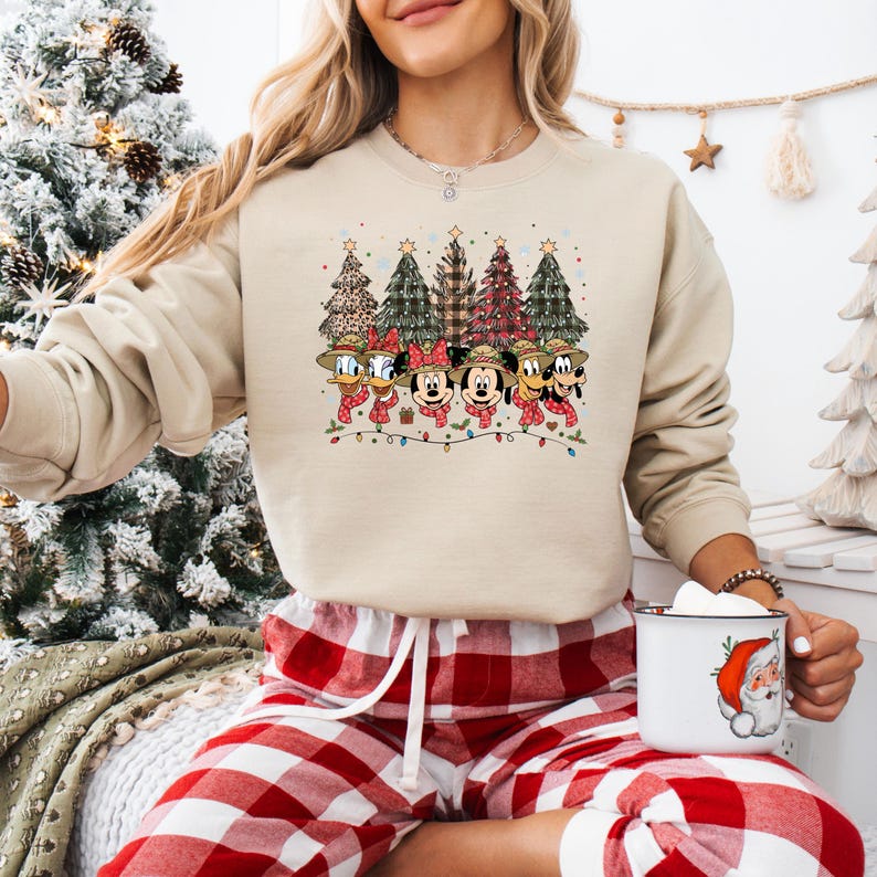 May include: Beige crewneck sweatshirt featuring a festive design with Disney characters, including Mickey and Minnie Mouse, Donald Duck, and Goofy, wearing scarves and hats. Christmas trees and string lights complete the holiday theme.