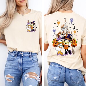 May include: Beige t-shirt featuring a Halloween-themed graphic of Mickey and Daisy Duck in witch costumes. The front has a small image, while the back showcases a larger design with a castle, pumpkins, and bats.