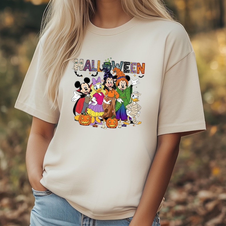 May include: Beige t-shirt featuring a Halloween graphic with Disney characters dressed in costumes. The word "HALLOWEEN" is printed above the characters. Mickey Mouse, Minnie Mouse, Donald Duck, and other characters are depicted.