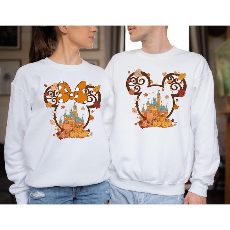 May include: A white sweatshirt with a brown Mickey Mouse head design featuring a fall-themed Cinderella's Castle and orange pumpkins. The design also includes a red and white polka dot bow.