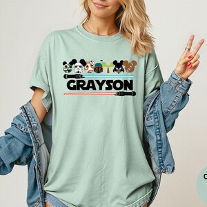 May include: A light green t-shirt with a graphic featuring cartoon characters and lightsabers. The name "GRAYSON" is printed in bold black letters. The shirt is part of the Comfort Colors Bay collection.