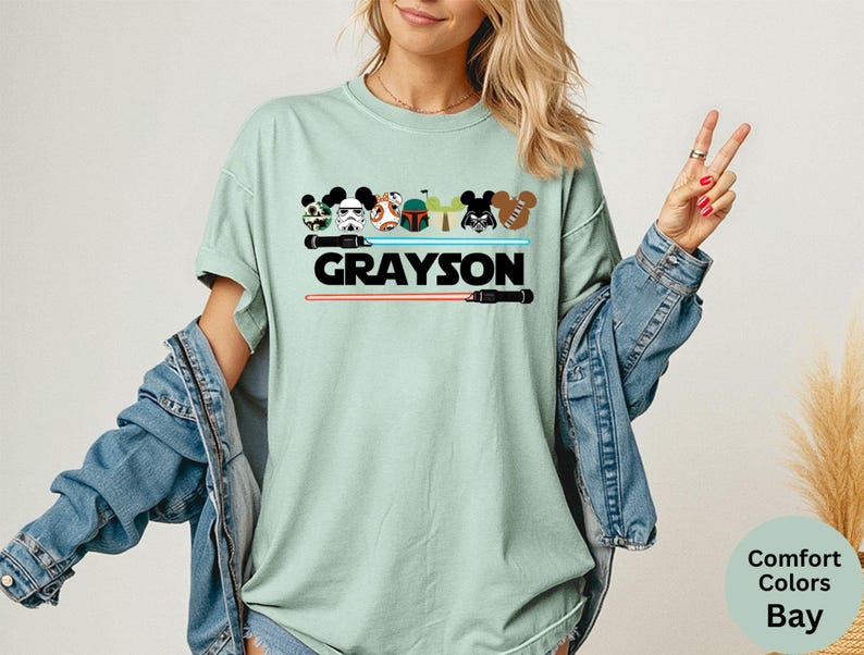 May include: A light green t-shirt with a graphic featuring cartoon characters and lightsabers. The name "GRAYSON" is printed in bold black letters. The shirt is part of the Comfort Colors Bay collection.