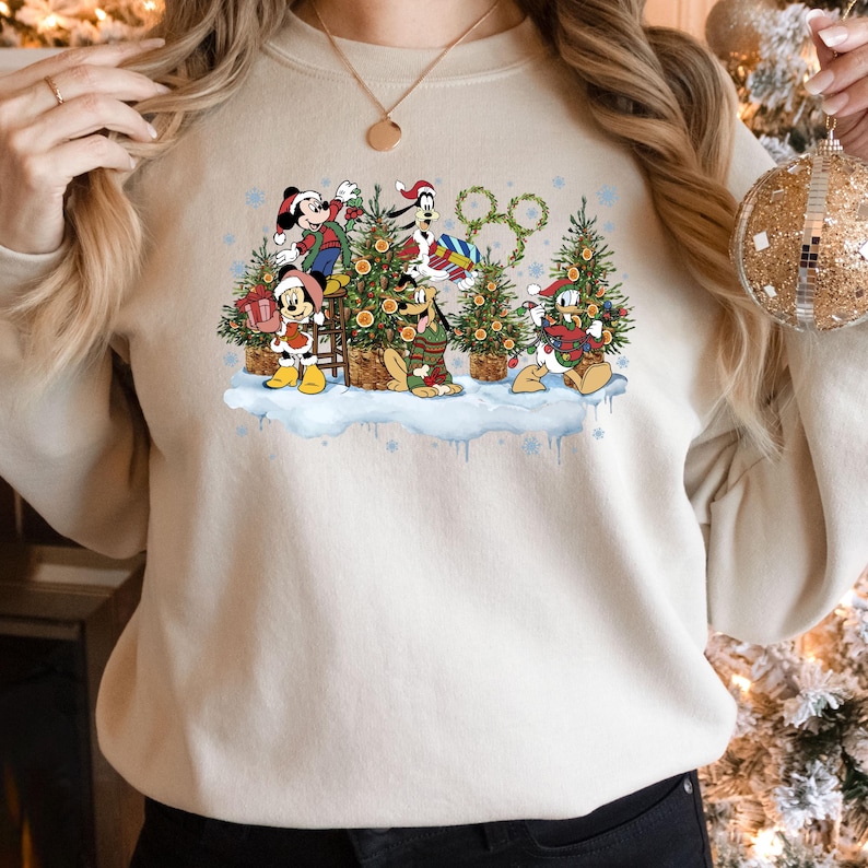 May include: A cream-colored sweatshirt with a Christmas-themed design featuring Mickey Mouse, Minnie Mouse, Goofy, Pluto, and Donald Duck decorating Christmas trees. The design includes a snow-covered ground, snowflakes, and the iconic Disney logo.
