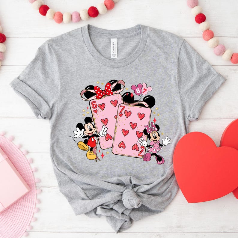 May include: A gray t-shirt featuring Mickey and Minnie Mouse with playing cards. The cards are pink with red hearts and the numbers 6 and 7. Mickey is wearing red shorts and Minnie is in a pink dress. A red bow is on Minnie's head.