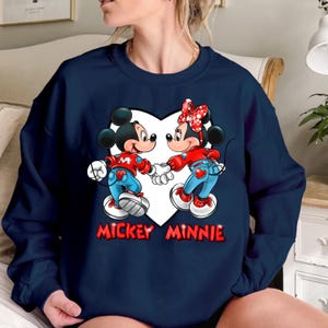 May include: A navy blue sweatshirt with a white heart graphic featuring Mickey Mouse and Minnie Mouse in red and blue outfits. The text "Mickey Minnie" is printed below the heart.
