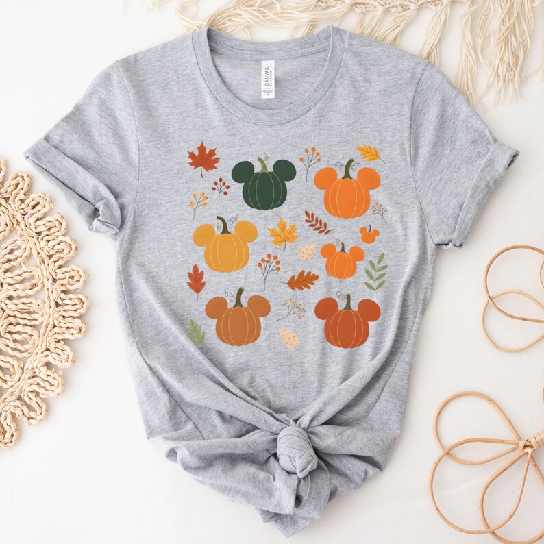 May include: A light gray t-shirt featuring a design of pumpkins with mouse ear shapes in various shades of orange and green. The design includes fall leaves in orange, red, and yellow.