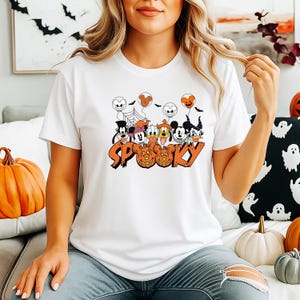 May include: White t-shirt with a Halloween graphic. The design features cartoon characters, balloons, and the word "SPOOKY" in orange. The shirt has a crew neck.