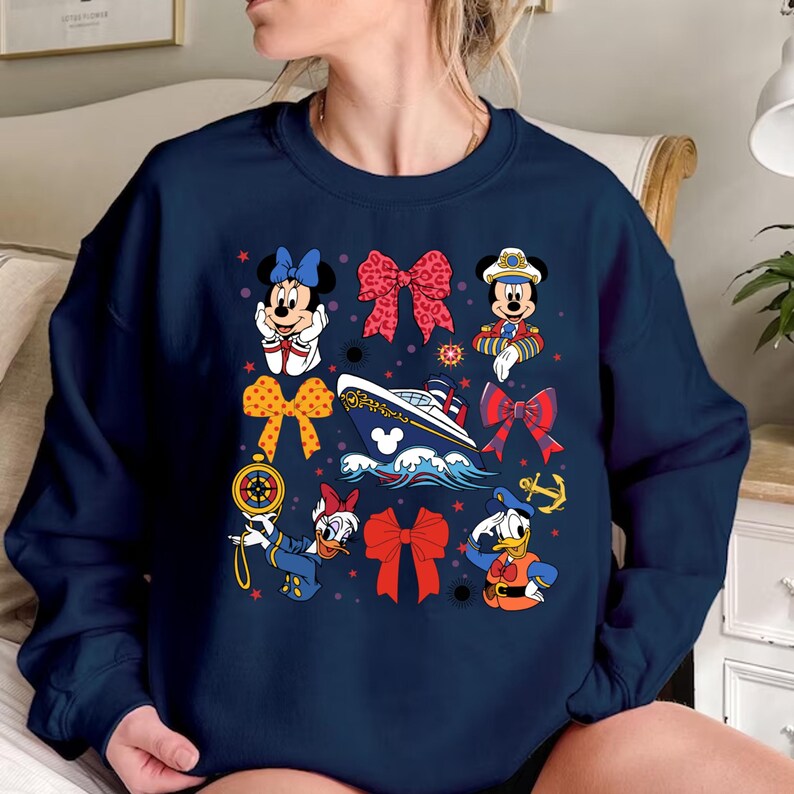 以下が含まれることがあります： ディズニーキャラクターがカラフルにプリントされたネイビーブルーのスウェットシャツ。ミッキーマウス、ミニーマウス、ドナルドダック、グーフィーなど。キャラクターは海事テーマの衣装を着ており、赤と白のリボン、船、その他の海事要素に囲まれています。