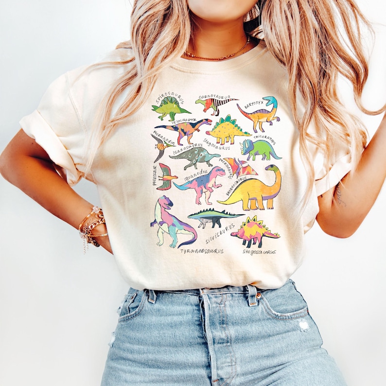 May include: A white t-shirt with a colorful graphic print of various dinosaurs. The dinosaurs are arranged in a circle with their names written underneath. The names include: Stegosaurus, Triceratops, Brachiosaurus, Tyrannosaurus, Iguanodon, Parasaurolophus, Corythosaurus, Pachycephalosaurus, Protoceratops, Ankylosaurus,  and more.