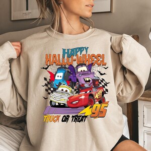 Może przedstawiać: Beżowa bluza z nadrukiem graficznym przedstawiającym kresk&oacute;wkowe samochody. Na bluzie nadrukowany jest tekst "Happy Hallo-Wheel Truck or Treat 95".