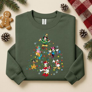 K&ouml;nnte beinhalten: Gr&uuml;nes Sweatshirt mit einem Weihnachtsbaum-Design aus Disney-Figuren, darunter Mickey und Minnie Mouse sowie Donald Duck. Das Design umfasst bunte Ornamente und einen Stern. Das Sweatshirt hat einen Rundhalsausschnitt und lange &Auml;rmel.