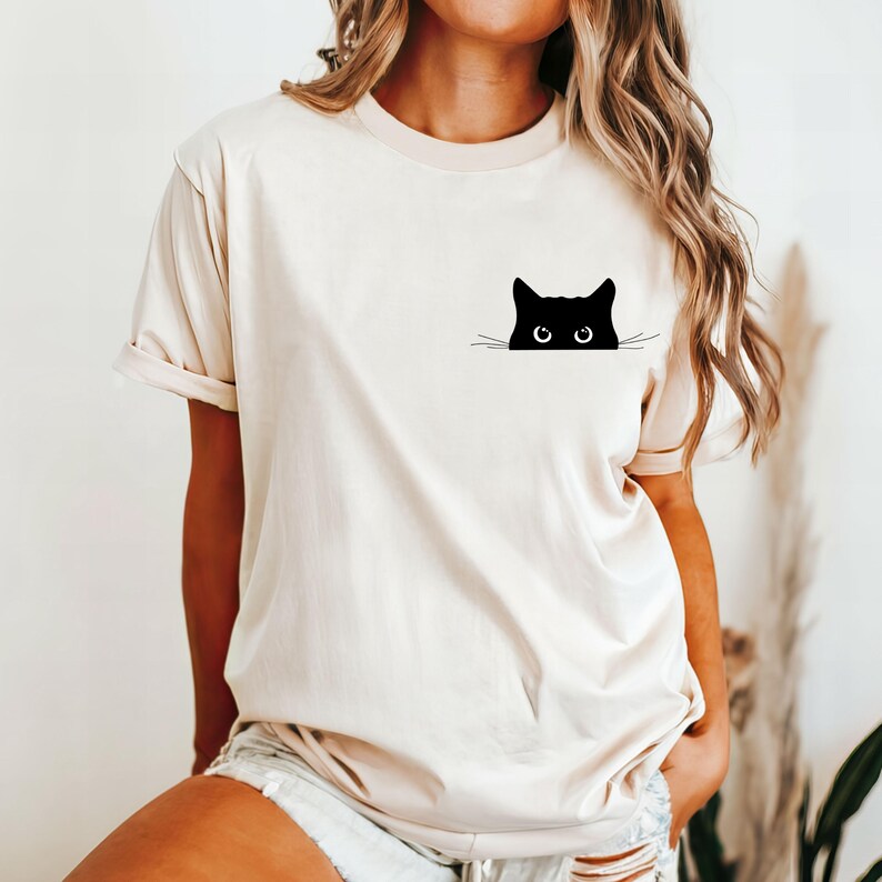 Pu&ograve; includere: Una maglietta bianca con un gatto nero che spunta da dietro il tessuto. Il gatto ha grandi occhi rotondi e un design semplice e minimalista.