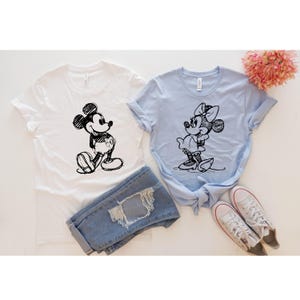May include: Two t-shirts with a sketch of Mickey Mouse and Minnie Mouse on them. The Mickey Mouse shirt is white and the Minnie Mouse shirt is light blue.