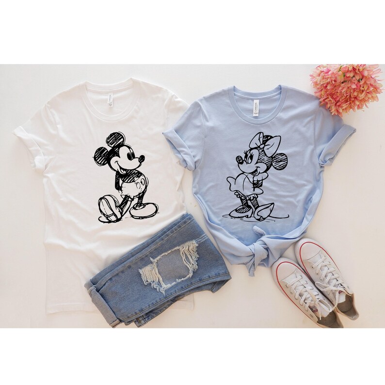 May include: Two t-shirts with a sketch of Mickey Mouse and Minnie Mouse on them. The Mickey Mouse shirt is white and the Minnie Mouse shirt is light blue.