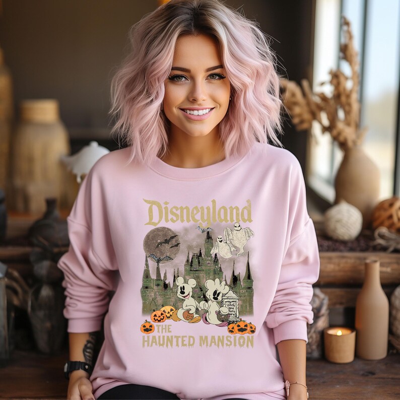 May include: A pink sweatshirt with a vintage-style graphic of Mickey Mouse and friends at the Haunted Mansion in Disneyland. The text "Disneyland" and "The Haunted Mansion" are printed on the shirt.