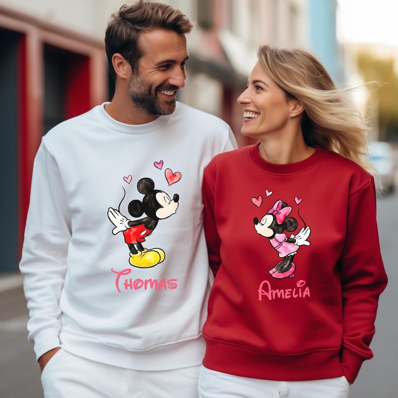 May include: Matching white and red sweatshirts featuring Mickey and Minnie Mouse graphics with hearts. The white sweatshirt has the name "Thomas" and the red sweatshirt has the name "Amelia". The sweatshirts are a couple's matching set.