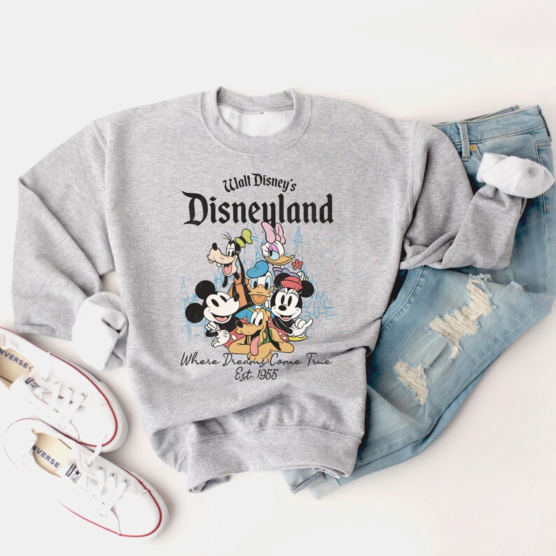 May include: A gray sweatshirt featuring the text "Walt Disney's Disneyland" above a graphic of Mickey Mouse, Donald Duck, Goofy, and other Disney characters. The sweatshirt is paired with blue jeans and white sneakers.