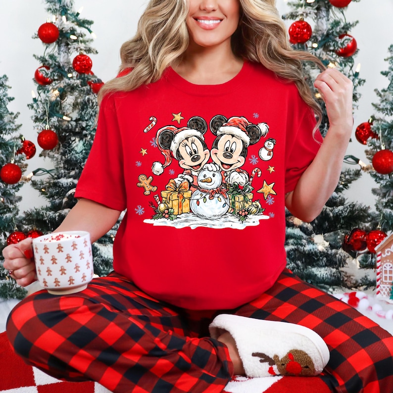 May include: A red short-sleeve shirt featuring Mickey and Minnie Mouse in Santa hats, with a snowman and gifts. The person is wearing red and black plaid pajama pants and white slippers. Christmas trees with red ornaments are in the background.