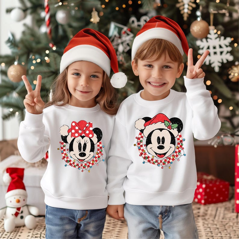May include: Two children wearing white Christmas sweaters with a Mickey Mouse and Minnie Mouse design. The sweaters feature a red and white checkered wreath with Christmas lights and holly. The children are wearing red Santa hats.