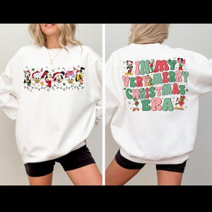 May include: White sweatshirt with a Christmas-themed design featuring Mickey Mouse and other Disney characters. The front of the sweatshirt has a line of characters with Christmas lights and snowflakes. The back of the sweatshirt has the text "In My Very Merry Christmas Era" with characters around the text.