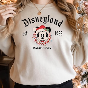 Puede incluir: Una sudadera blanca con un logotipo de Disneyland de estilo vintage que presenta a Mickey Mouse con un gorro de Pap&aacute; Noel y rodeado de luces navide&ntilde;as. El texto dice "Disneyland est 1955 California".