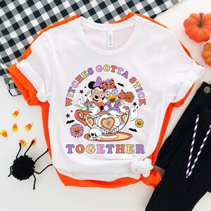 K&ouml;nnte beinhalten: Wei&szlig;es T-Shirt mit einem Aufdruck von Mickey Mouse und Minnie Mouse, die als Hexen in einer Teetasse verkleidet sind. Der Text "Witches Gotta Stick Together" ist unter dem Bild gedruckt.
