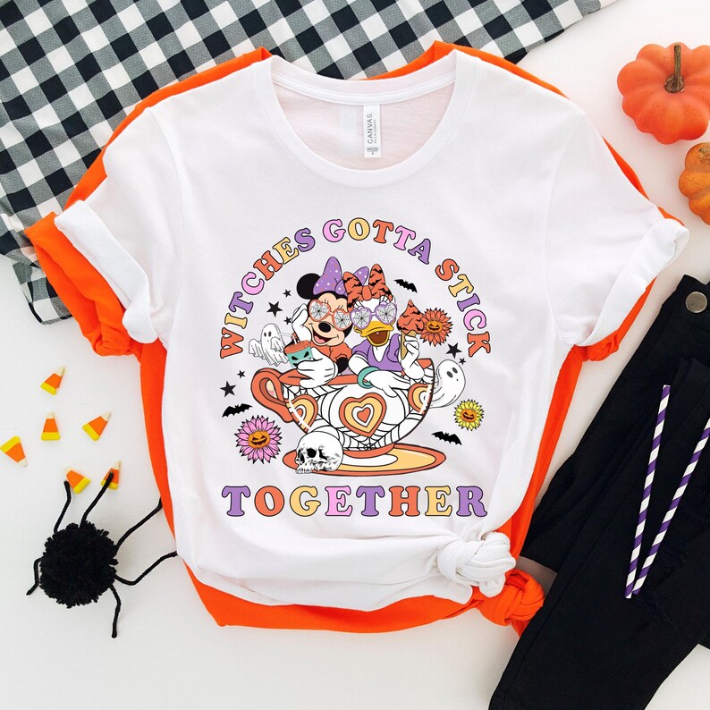 K&ouml;nnte beinhalten: Wei&szlig;es T-Shirt mit einem Aufdruck von Mickey Mouse und Minnie Mouse, die als Hexen in einer Teetasse verkleidet sind. Der Text "Witches Gotta Stick Together" ist unter dem Bild gedruckt.